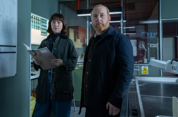 Vanessa Emme and Adrian Scarborough as Layla Walsh and Max Arnold. They're standing side-by-side in a laboratory; she's holding a clipboard and they're looking directly at the camera.