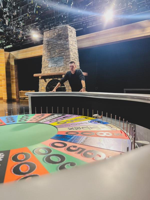 Ryan Seacrest looking at a large wheel. 
