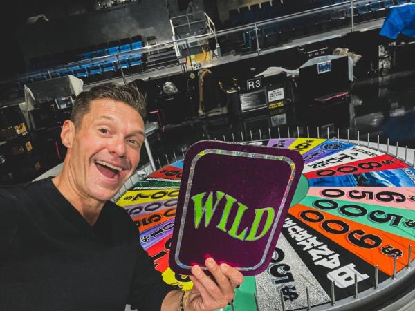 Ryan Seacrest smiling next to a wheel. 