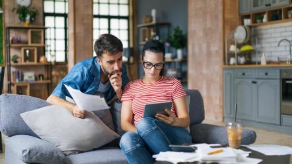 Couple calculating bills at home using tablet and calculator. Young couple working on computer while calculating finances sitting on couch. Young <em></em> man with <em></em> wife at home analyzing their finance with docu<em></em>ments.