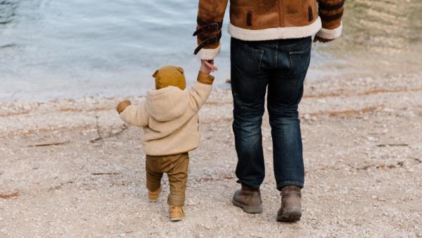 A man and a toddler in coats by a lake, holding hands and facing away from the camera.