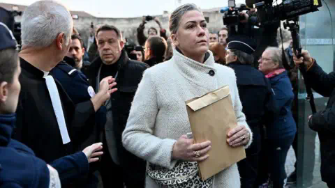 Getty Images Caroline Darian, wearing a cream coat, walks through a crowd, holding a brown envelope. She is followed by her brothers and there are cameras to her left.